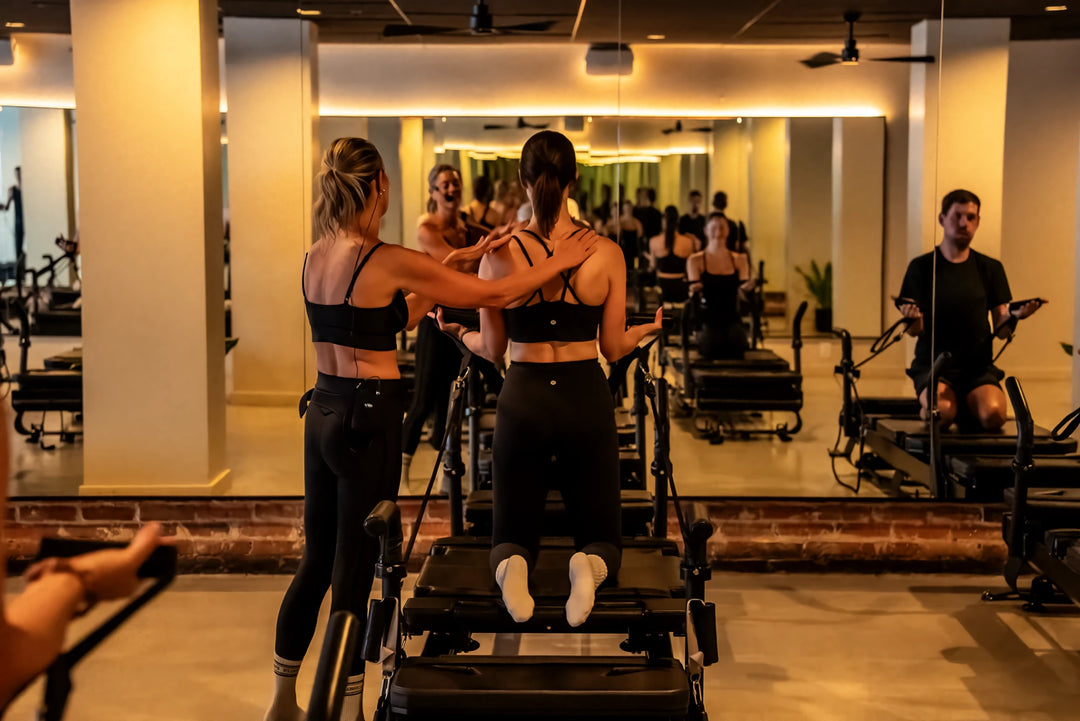 Pilates instructor with a headset mic adjusting a kneeling client's shoulders during a FlowCorps reformer class