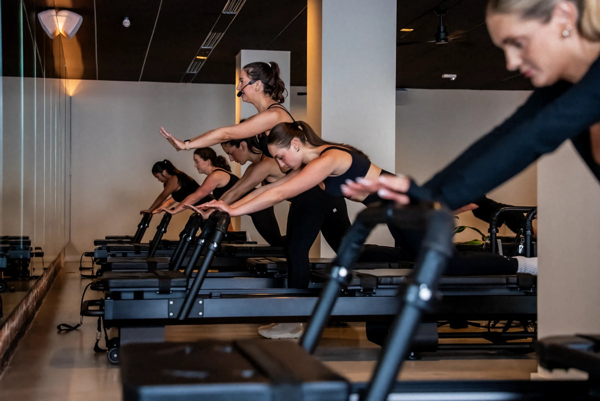 Instructor with a headset mic leading clients through a forward reach in a FlowCorps reformer Pilates class