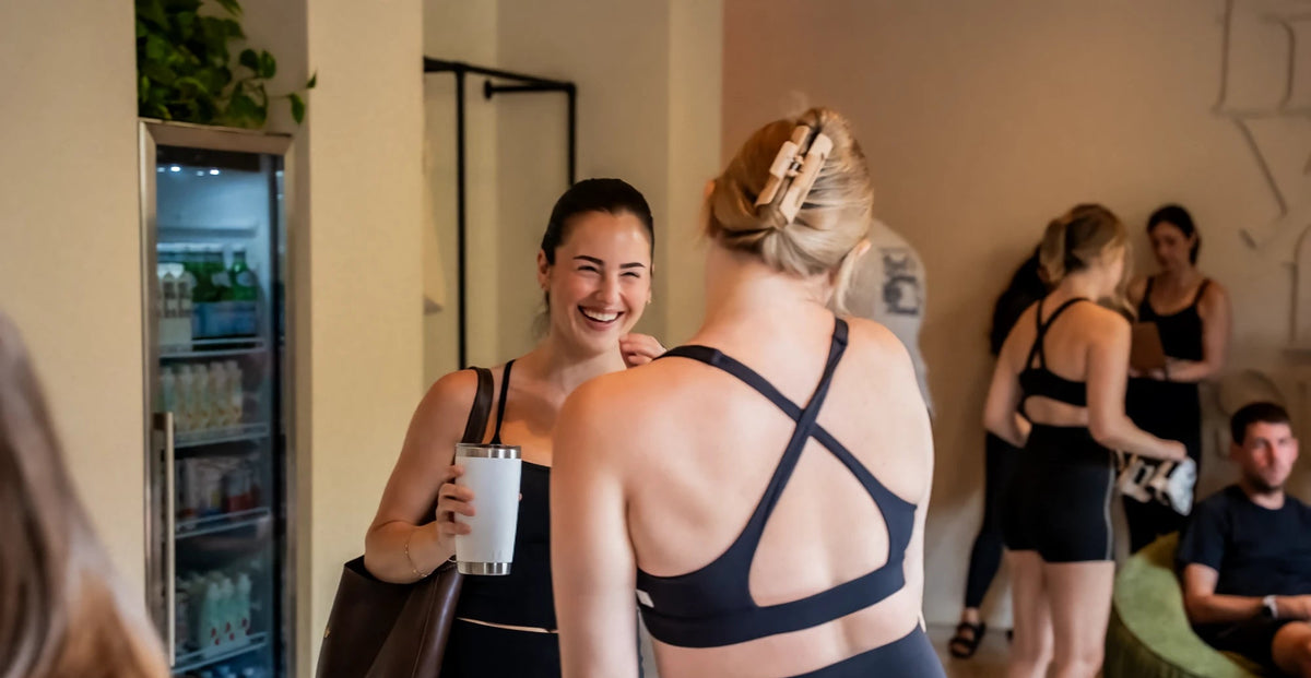Smiling client holding a tumbler and chatting with a friend in the FlowCorps studio lobby before class